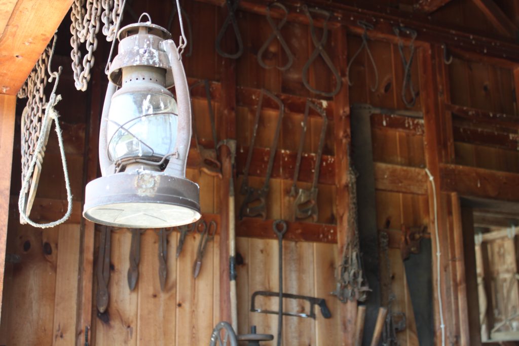 An old-timey lantern hanging in front of a wall of iron tools in a barn.