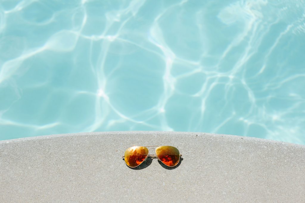 A pair of orange-lensed sunglasses sitting on a curved section of concrete next to a sun-lit swimming pool.
